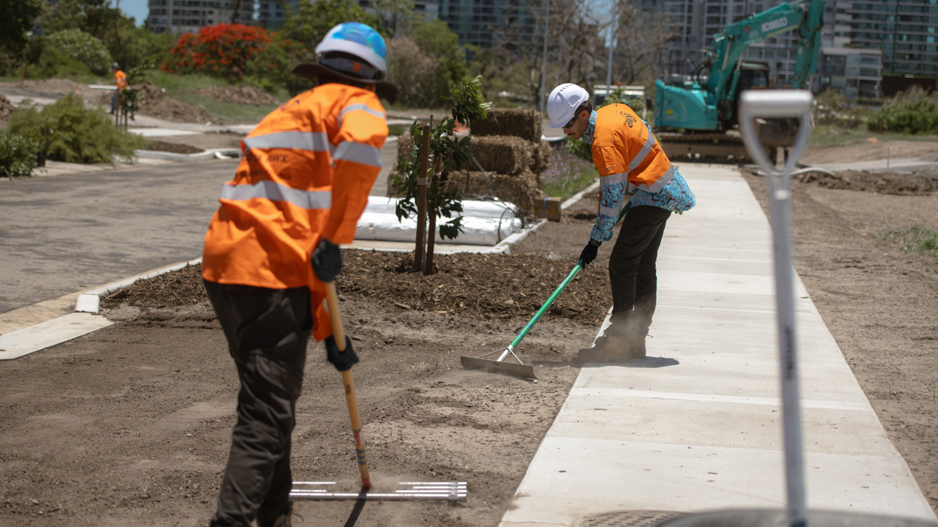 Two workers in orange vests and hard hats raking soil near a concrete path.