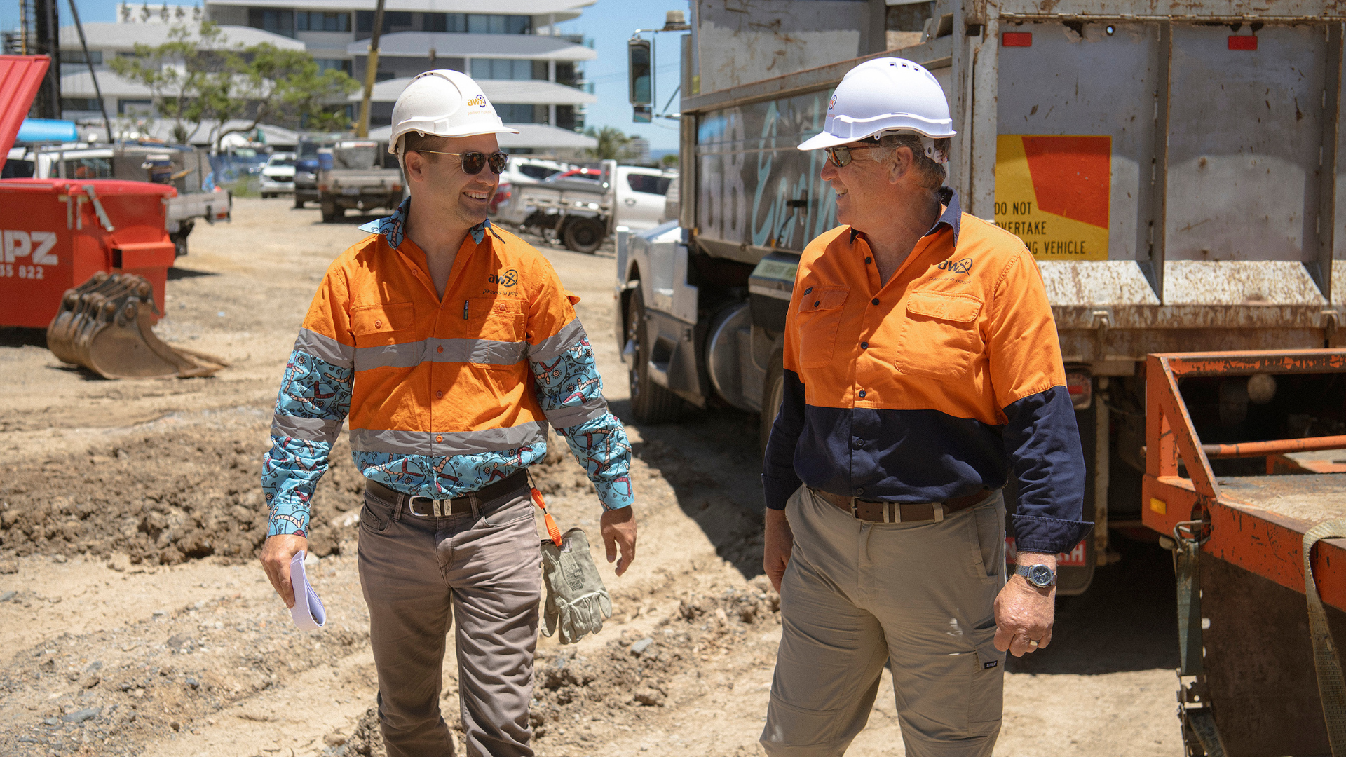 Two construction workers in safety vests and hard hats on a worksite, talking near a truck.