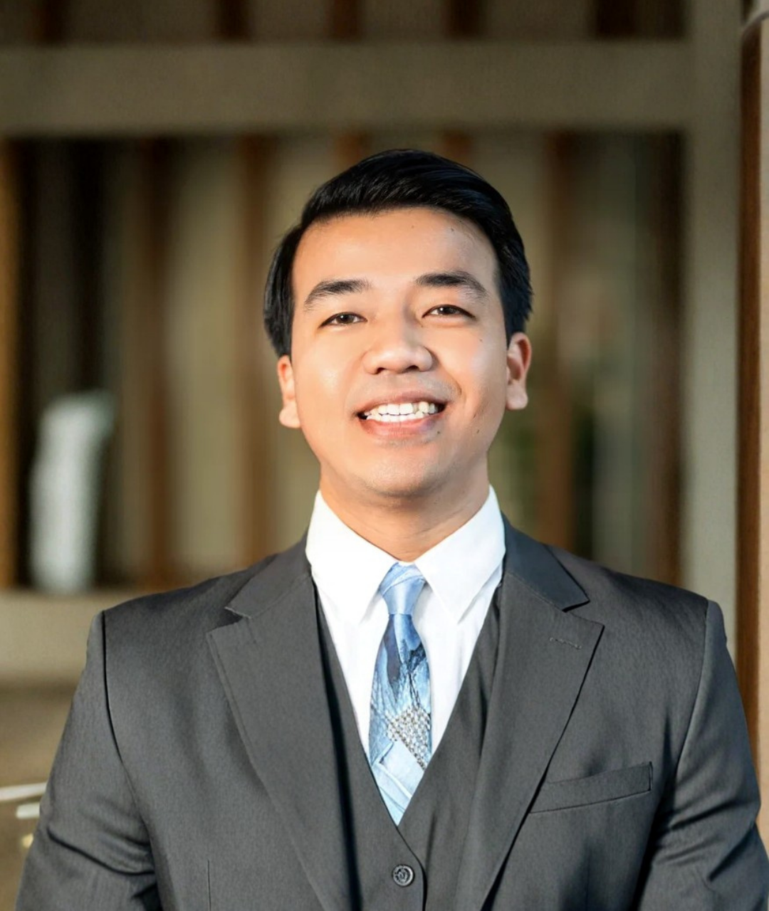 Man in orange and blue work shirt smiles, arms crossed, leaning against wooden post. Outdoors setting.