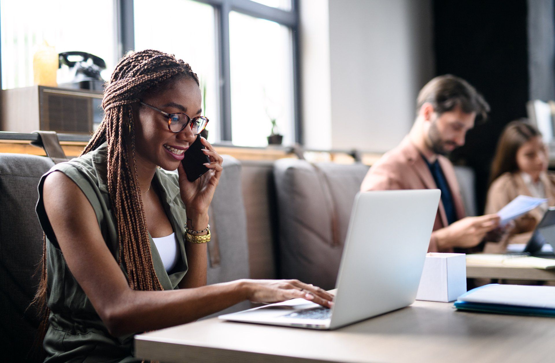 A woman is sitting at a table using a laptop and talking on a cell phone.