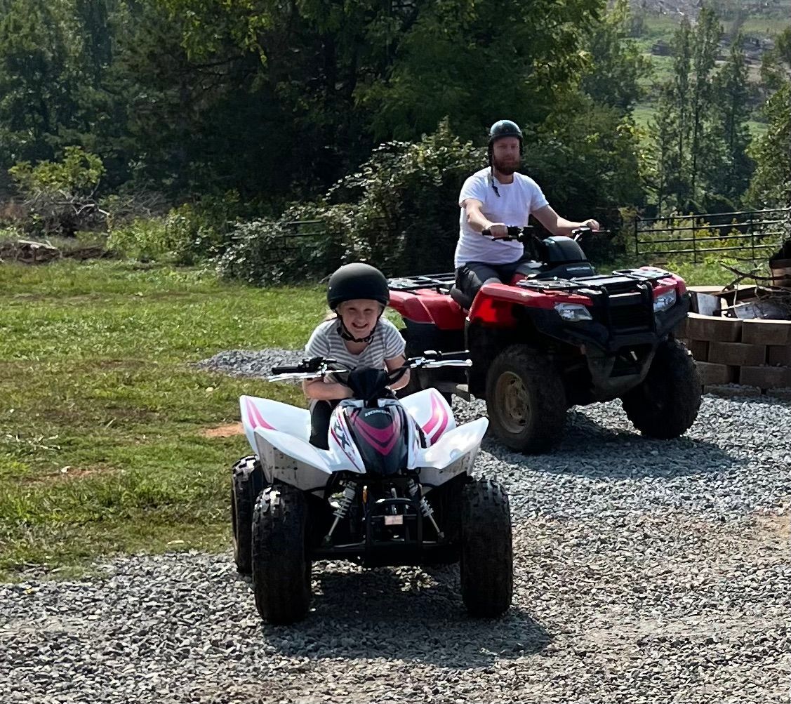 Child and man ride ATVs on gravel in a grassy area, wearing helmets; trees and building in background.