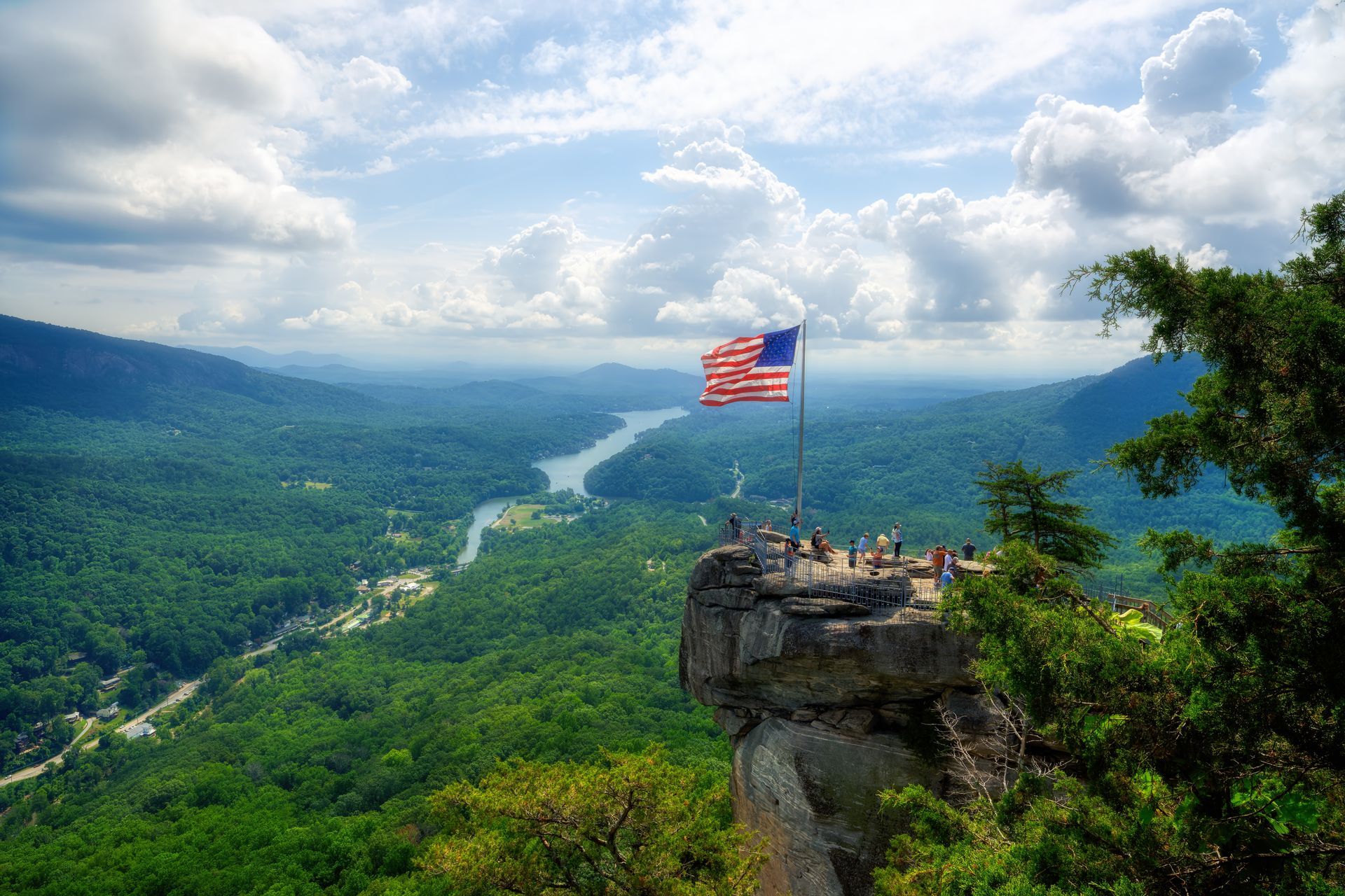 American flag waves atop a rocky cliff overlooking a lush, green valley and river.