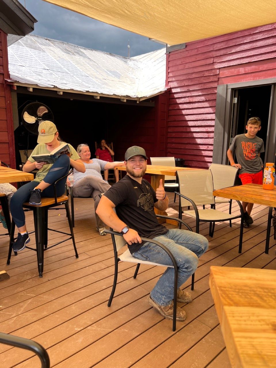 People seated at outdoor tables on a wooden deck, red building in the background. Man in the foreground gives a thumbs-up.