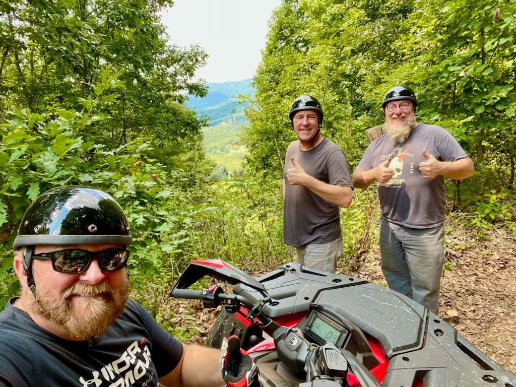 Three men with helmets pose with an ATV on a mountain trail, giving thumbs up.
