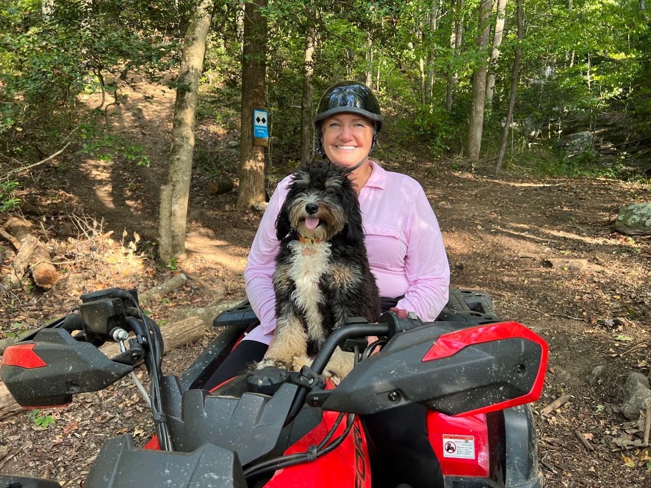 Woman and dog on a red ATV on a forest trail; both are smiling.