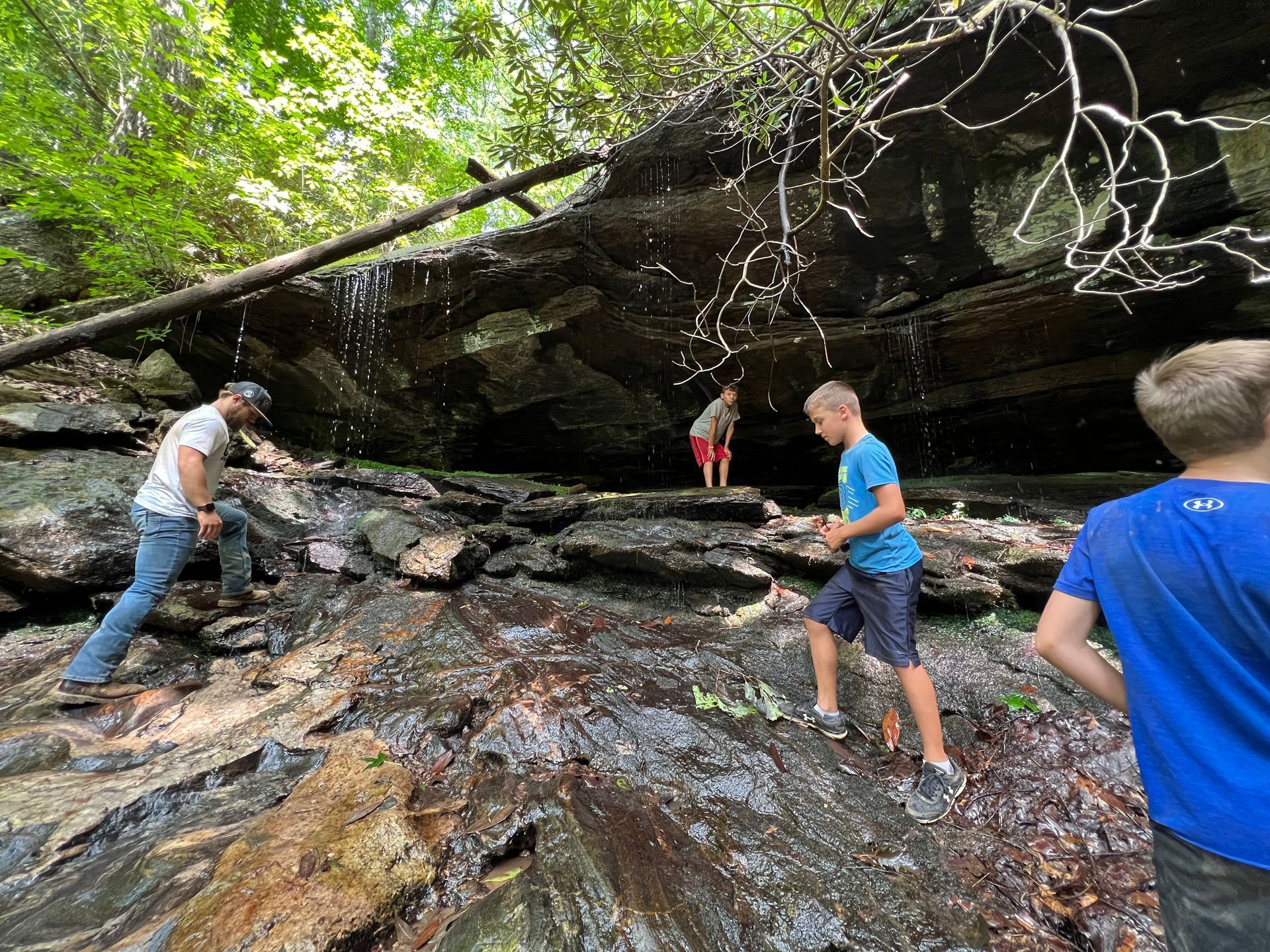 People exploring a rocky, wet overhang in a forest, water trickling down.