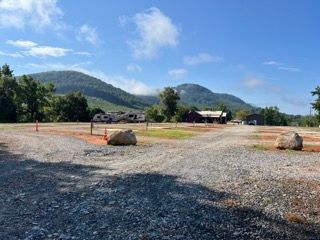 Gravel RV campsite with power hookups, a building, mountains, and a blue sky with clouds.