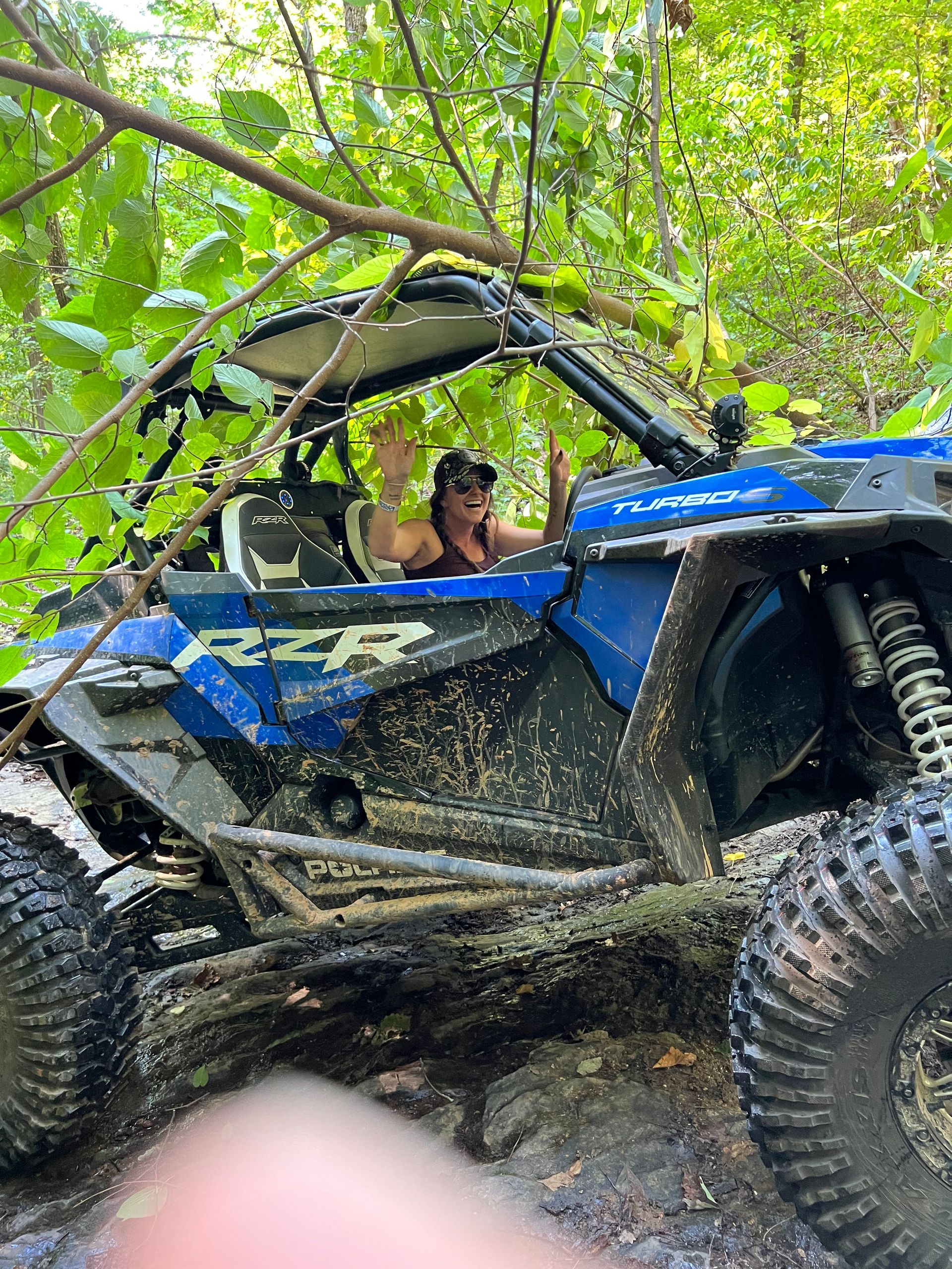 Person in blue off-road vehicle covered in mud, waving from a wooded area.