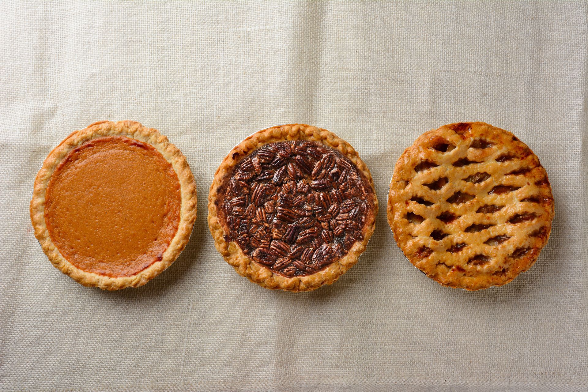 Three mini pies: pumpkin, pecan, and apple, on a beige cloth.