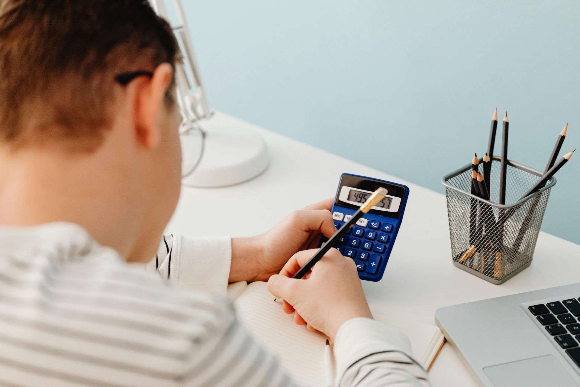 Person using a calculator and writing in a notebook at a desk with a laptop and pencils.