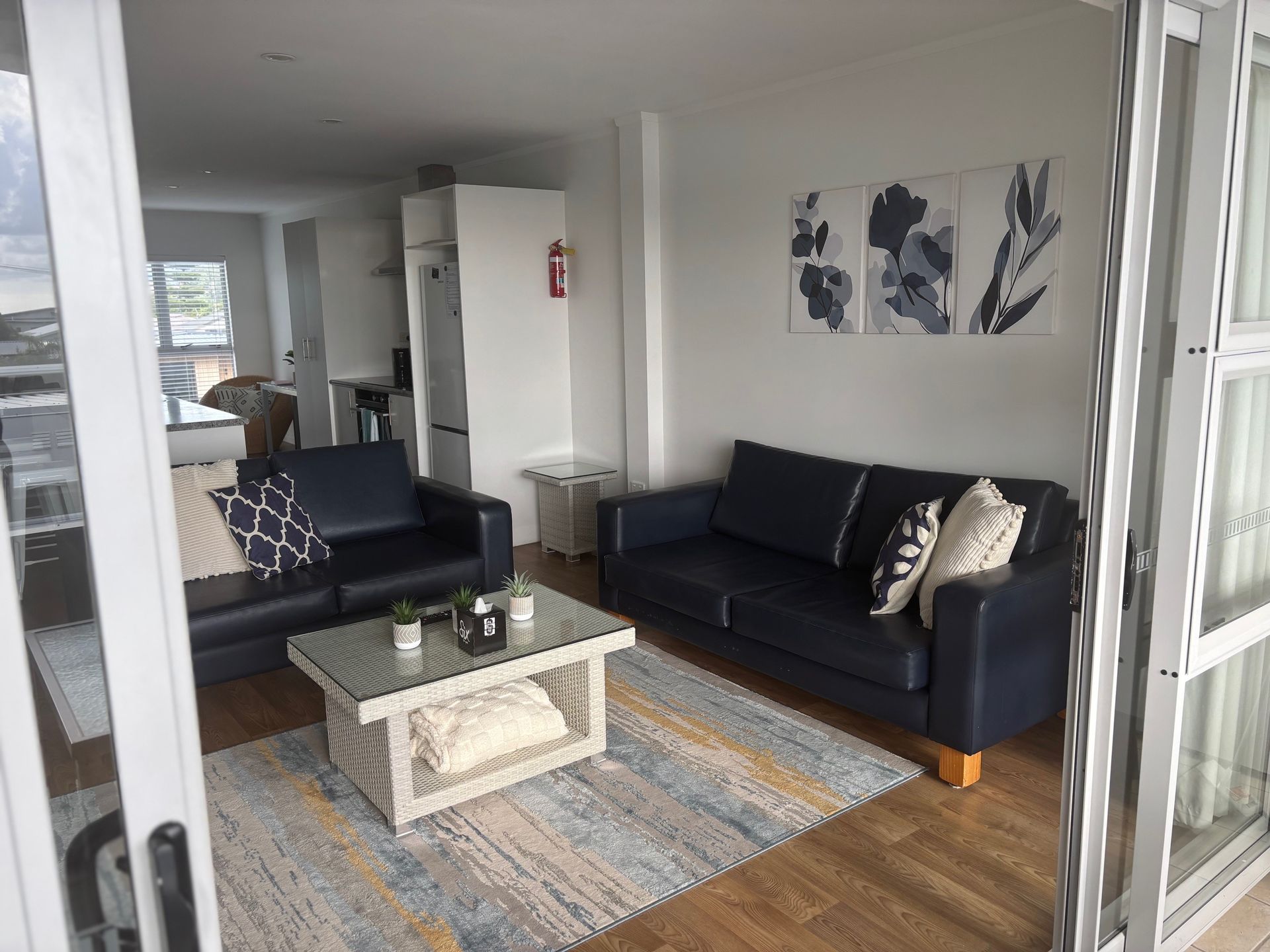Bright living room with two dark sofas, glass coffee table, blue rug, and wall art.