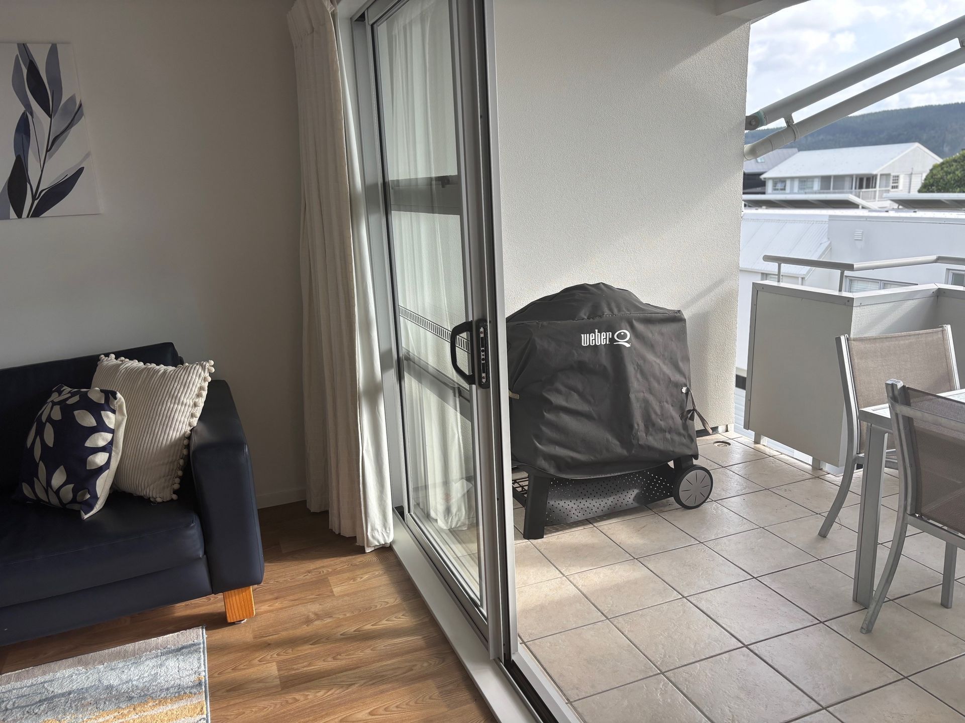 Living room with dark sofa opening to a tiled balcony with a covered grill and outdoor chairs