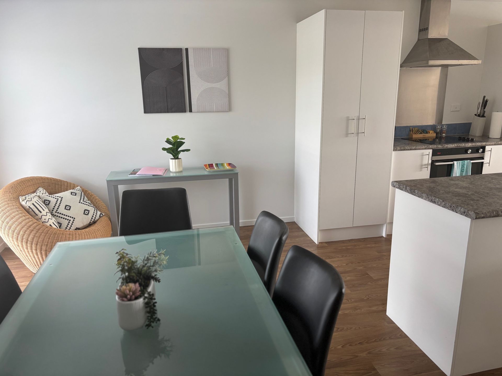 Dining and kitchen area with glass table, black chairs, white cabinets, and a wicker chair by the wall