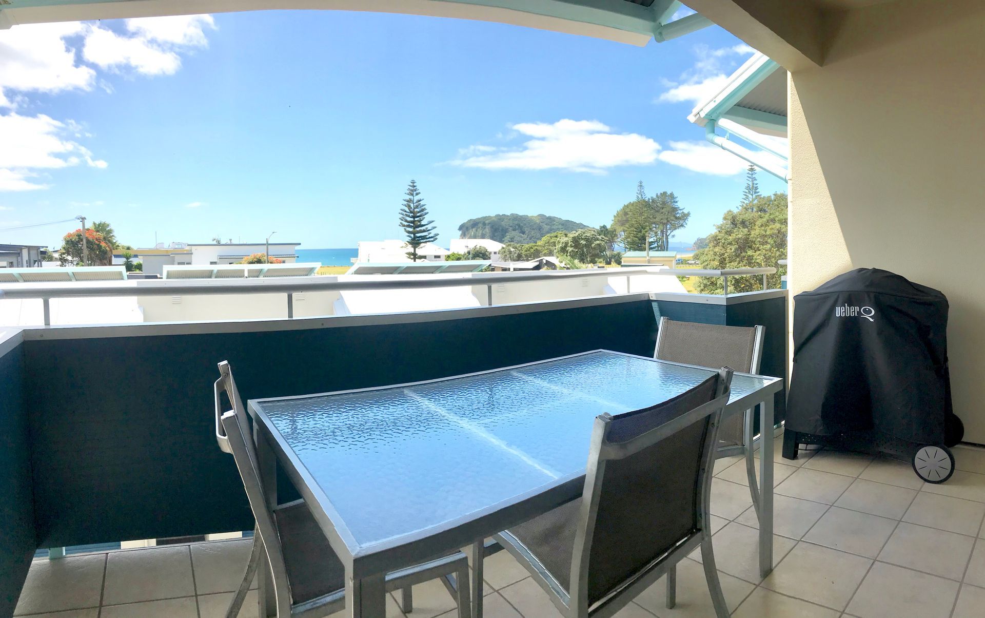 Balcony with glass-top table, four chairs, and a covered grill overlooking trees and rooftops under a blue sky