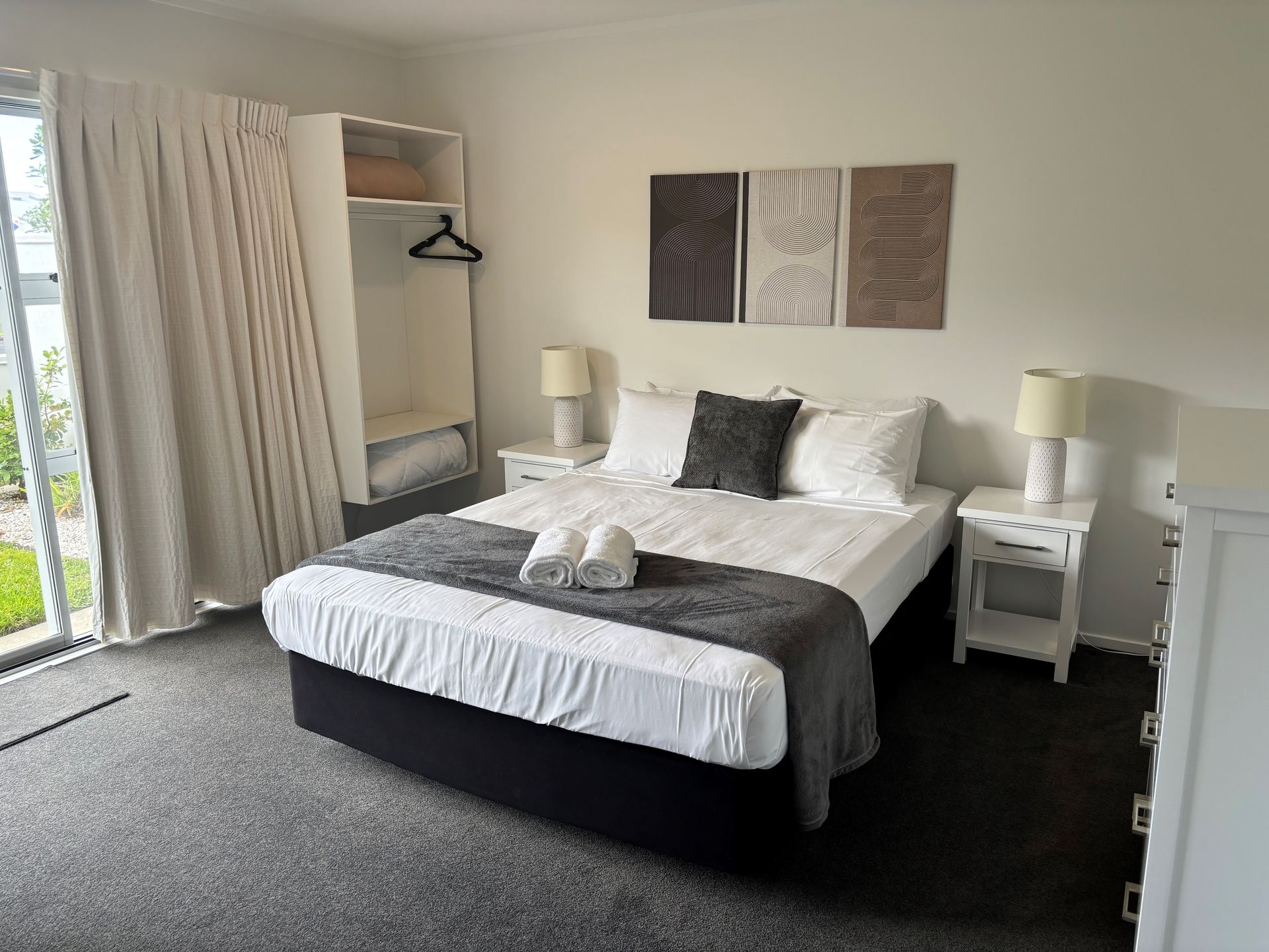 A hotel bedroom with a queen bed, grey throw, two nightstands with lamps, a closet, and wall art against white walls.