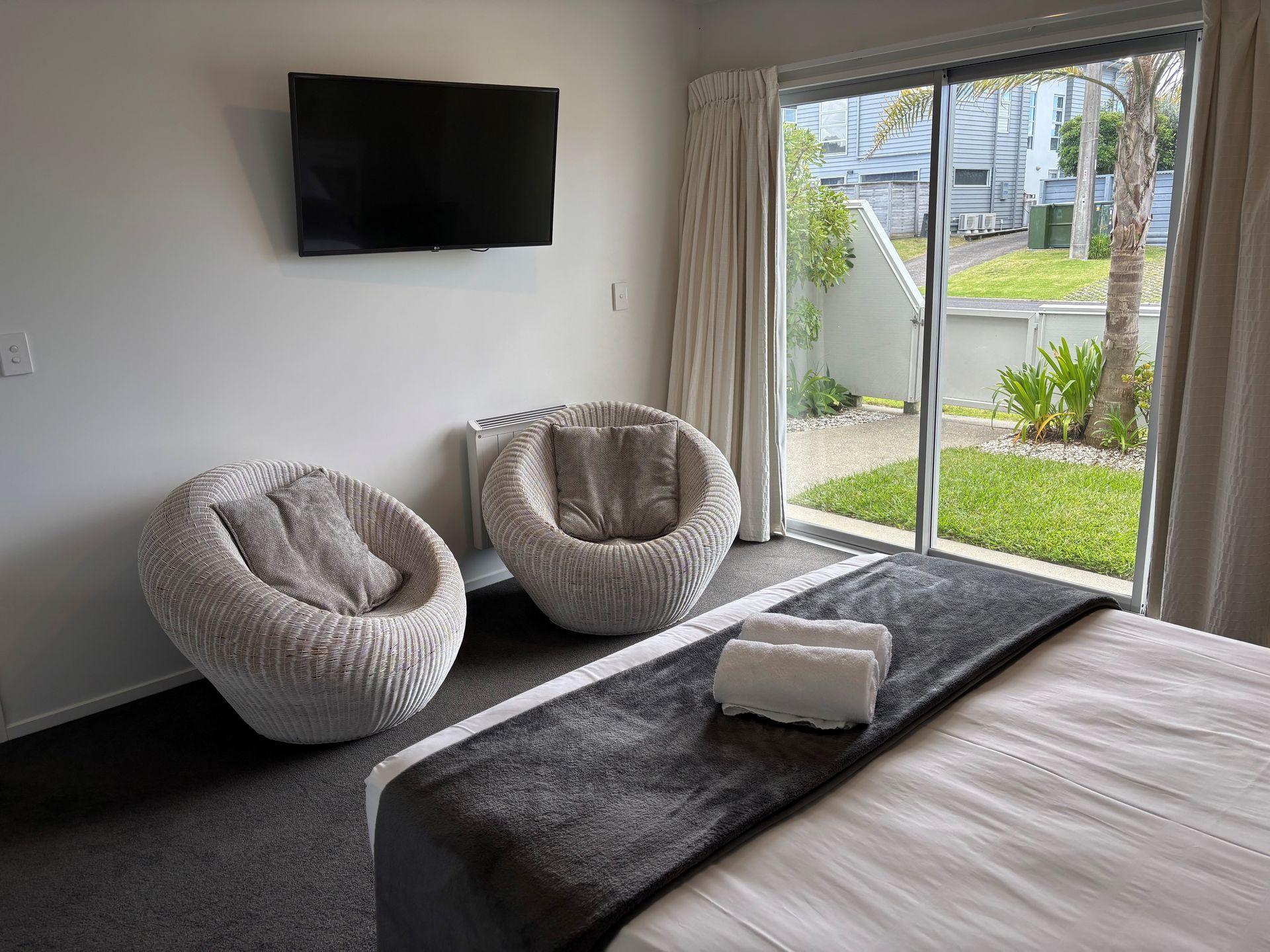 A modern hotel room featuring two round white wicker chairs, a gray bed runner, and a sliding glass door to a patio.