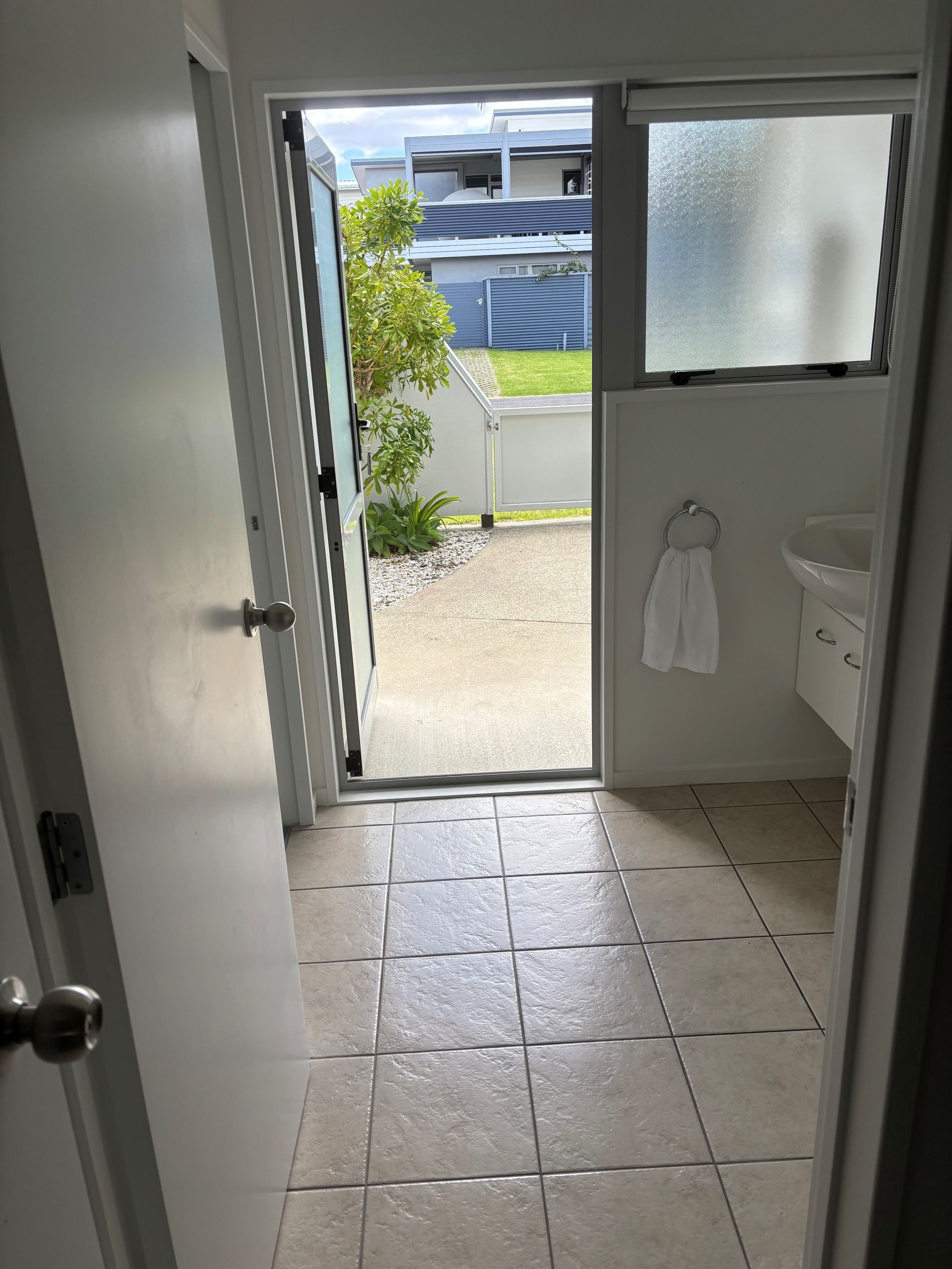 A view from a doorway into a tiled bathroom with a sink, leading to an open door revealing an outdoor gravel area.