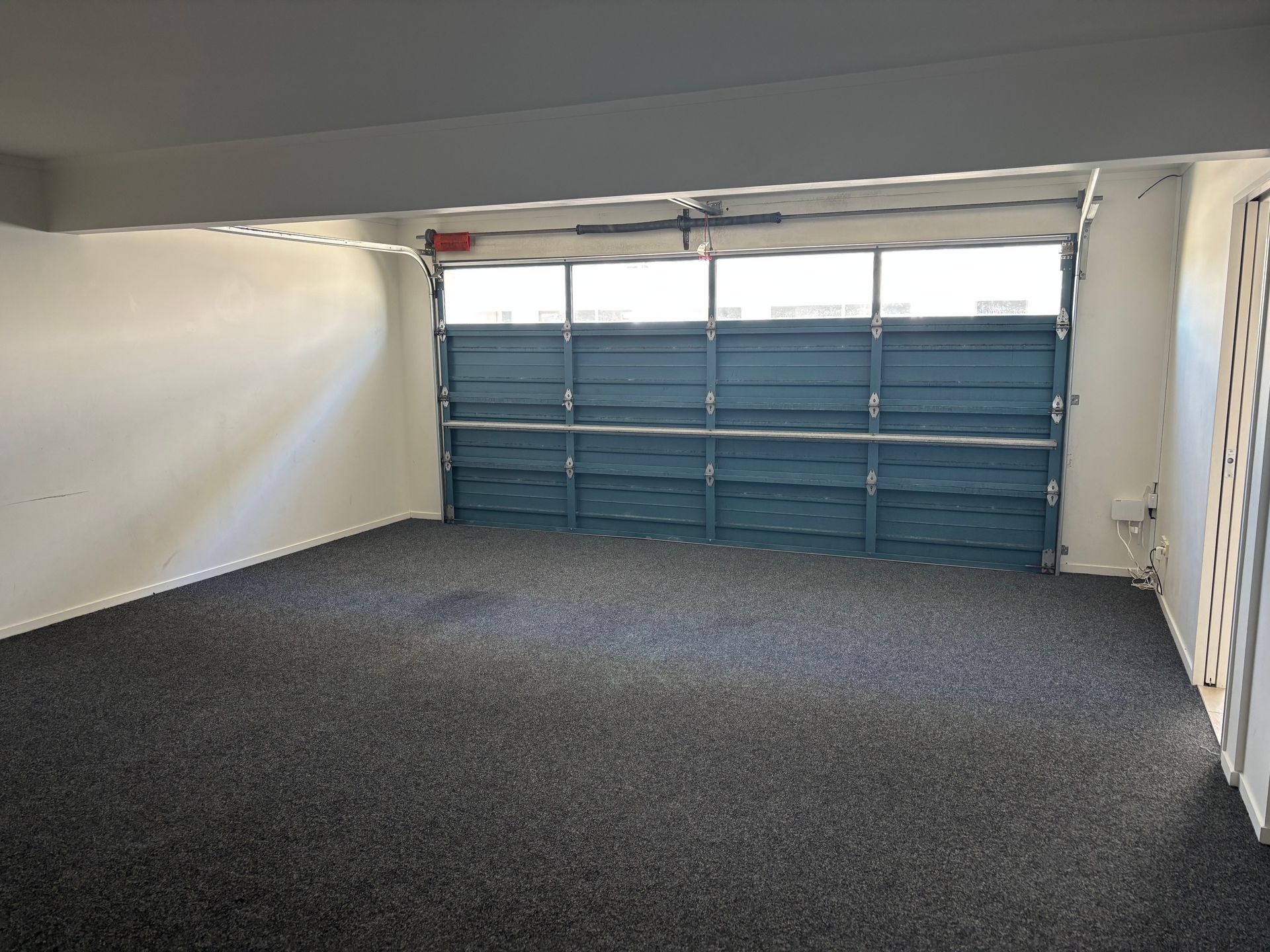 An empty garage with gray carpeted floors, white walls, and a blue paneled garage door.