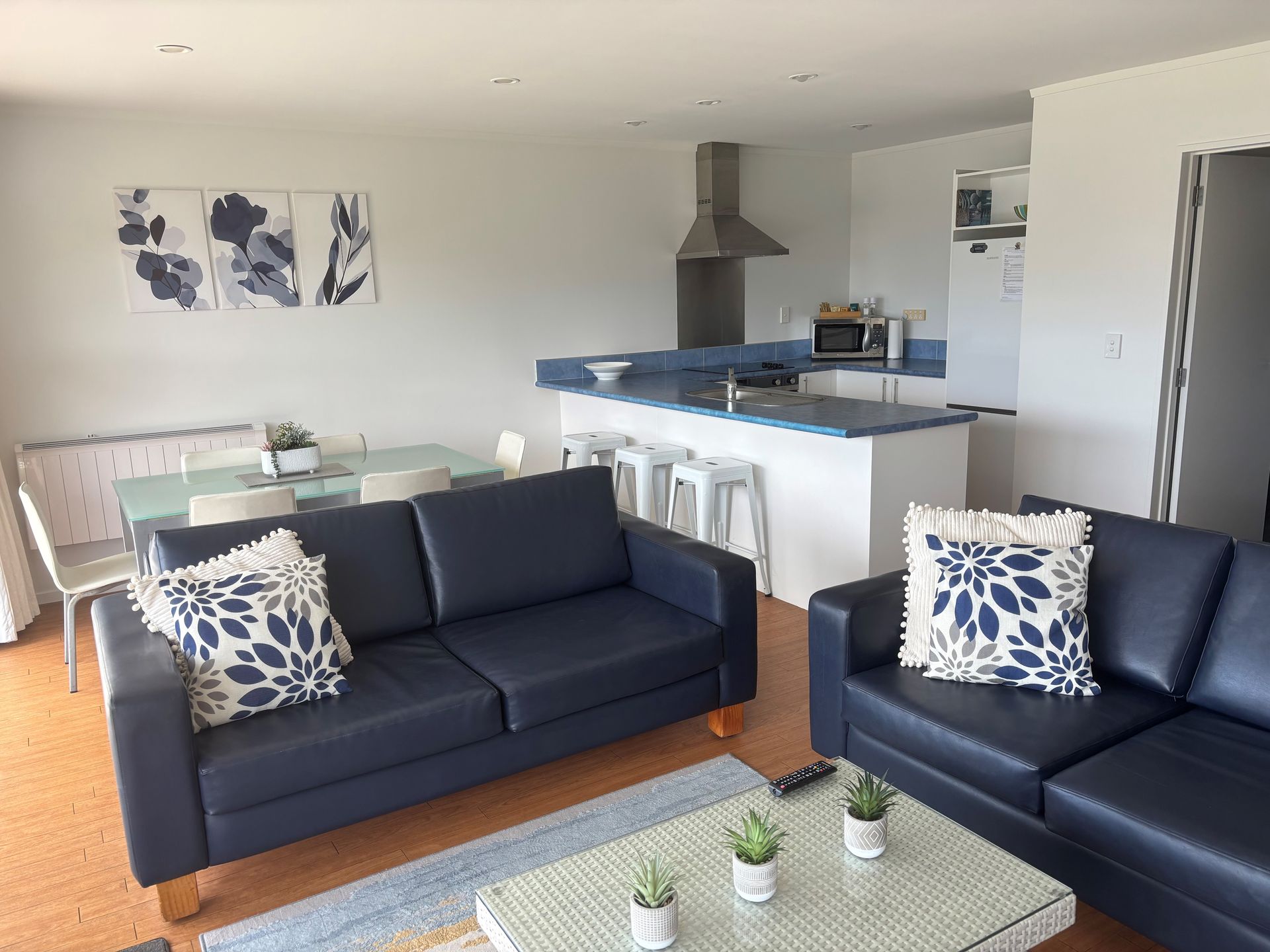 A living room and kitchen area featuring two dark navy blue sofas, a glass coffee table, and a kitchen island with stools.