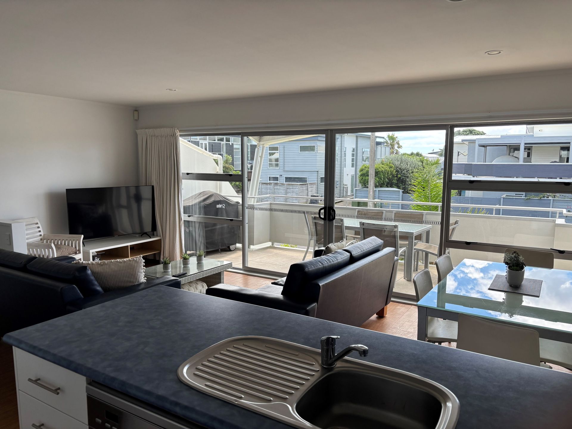 An open-plan kitchen looking into a living room with dark sofas, a television, and a deck with outdoor dining.