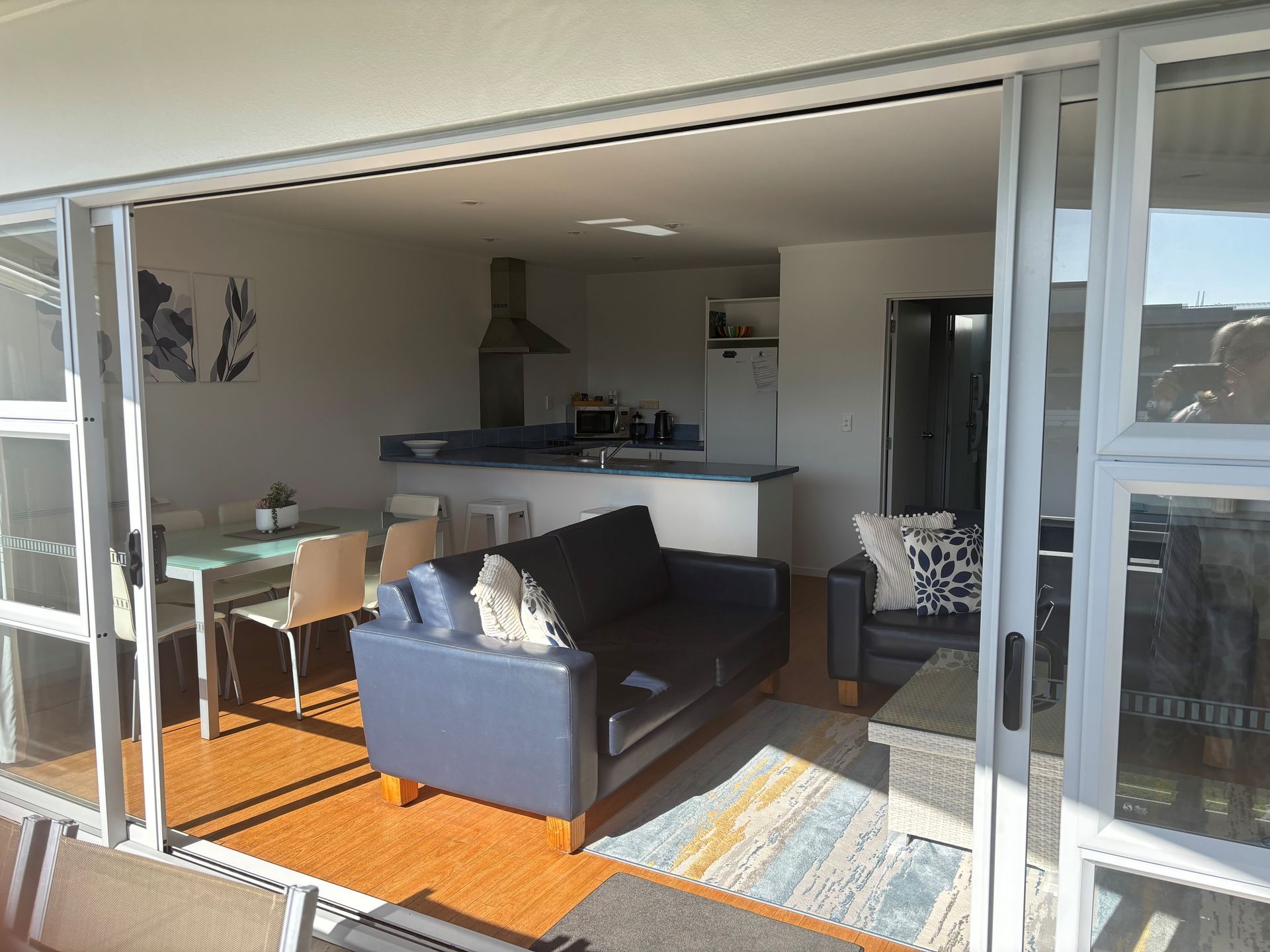 Sliding doors open to a sunlit living room with a black sofa, dining table, and kitchen.