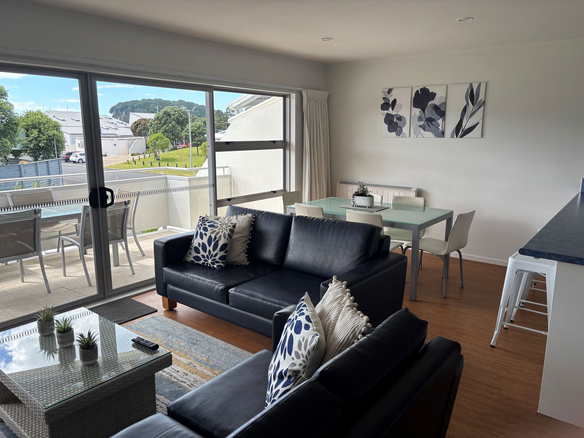A living room featuring black leather sofas, a glass-topped coffee table, and a dining area beside a large sliding door.