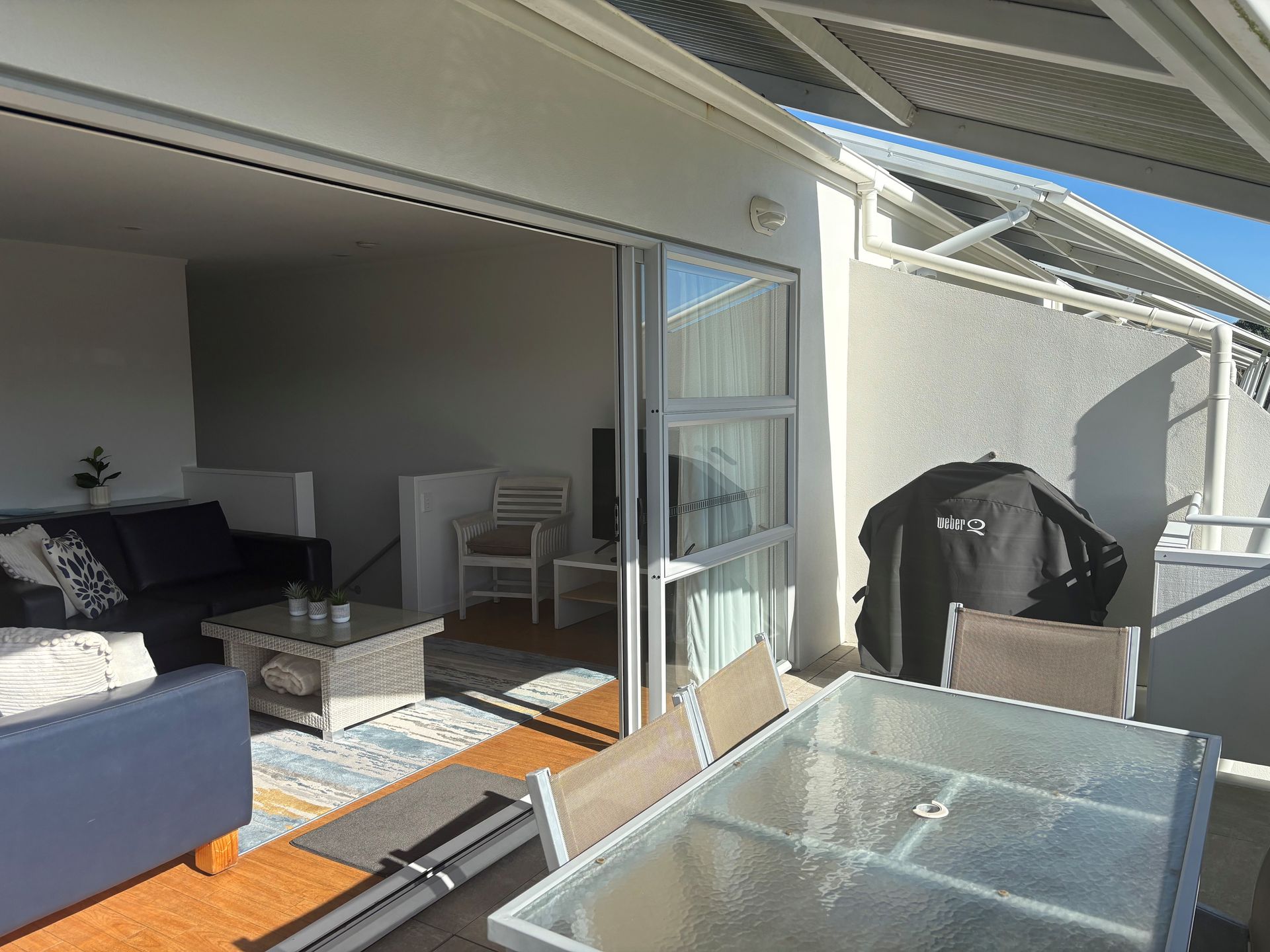 An indoor-outdoor living area featuring a blue sofa, a glass patio table, and a covered grill on a sunny balcony.