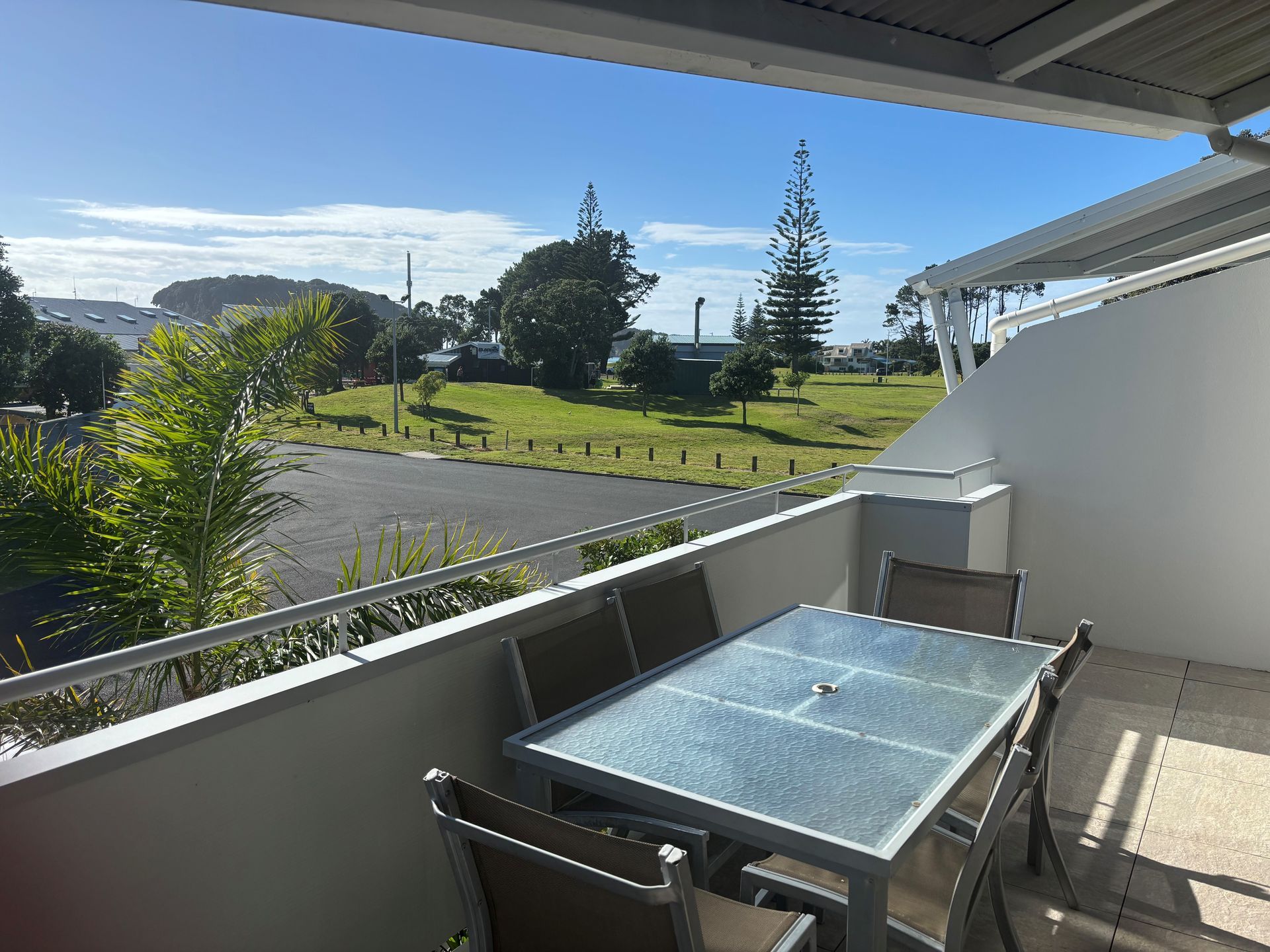 Balcony with glass table overlooking a sunny green park and road beyond.