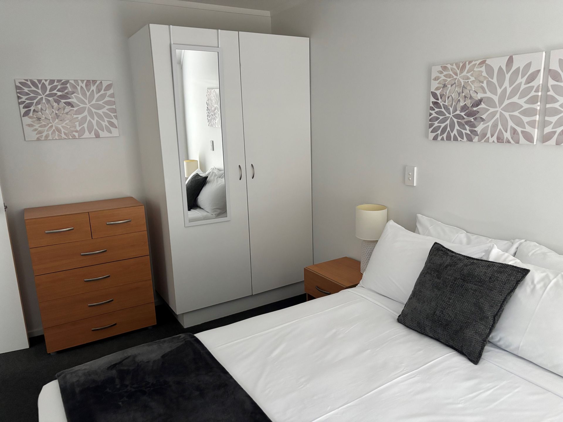 A bedroom with a white wardrobe, wooden chest of drawers, bed with white bedding, grey throw, and matching floral wall art.