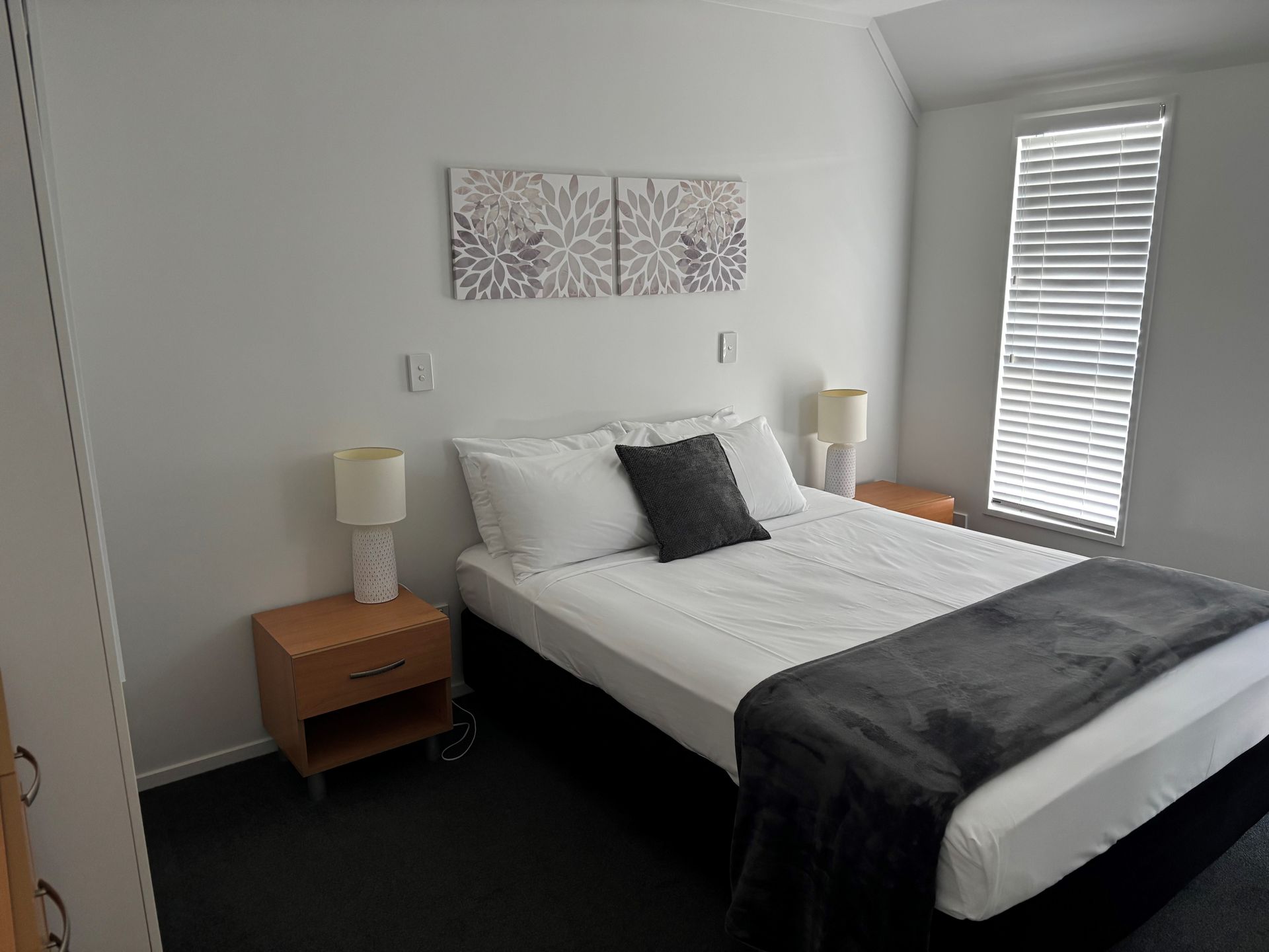 A bedroom with a bed featuring white linens, a dark gray runner, two nightstands with lamps, and floral wall art.