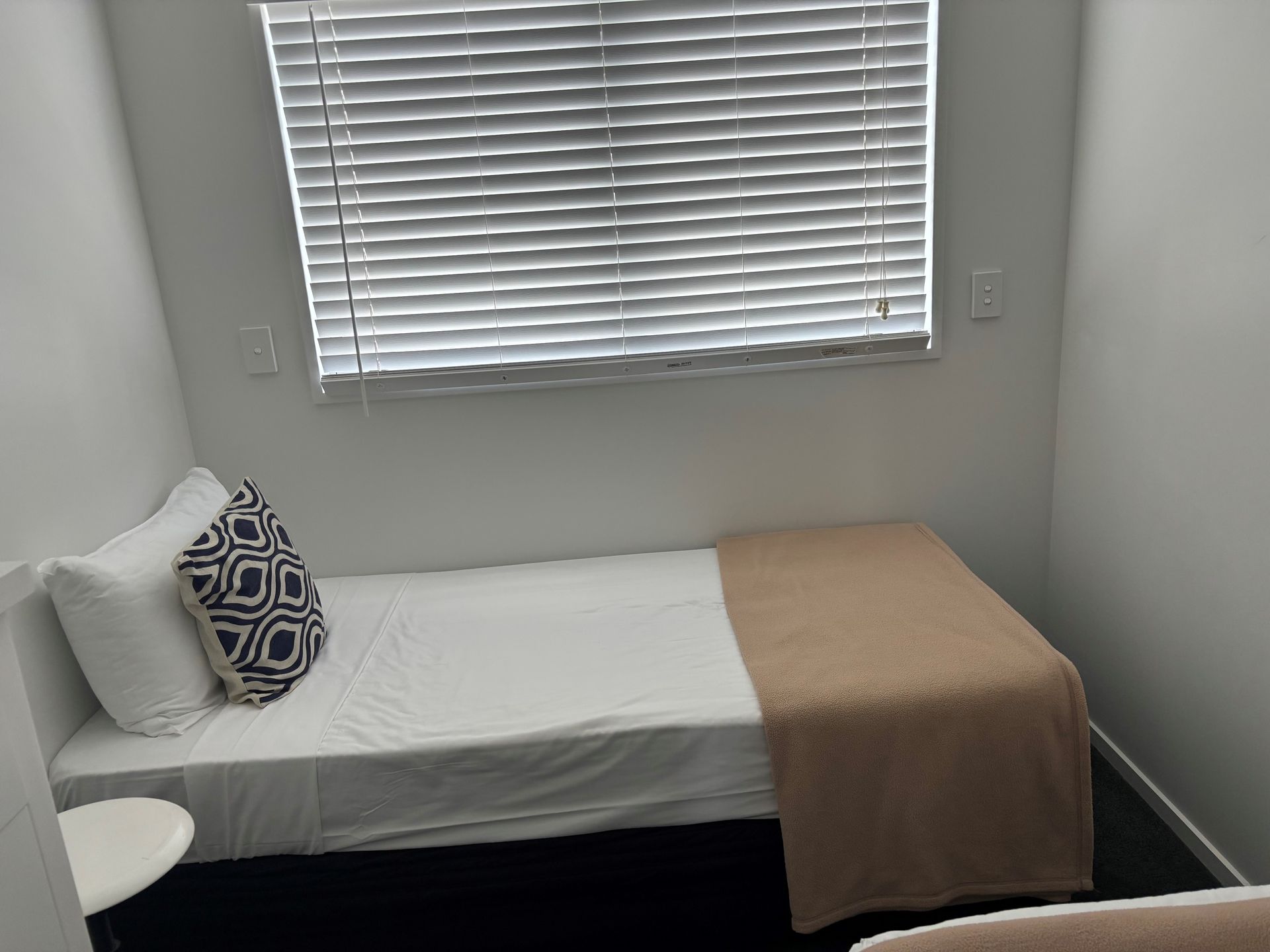 A single bed with white linens and a tan throw blanket, positioned under a window with closed horizontal white blinds.