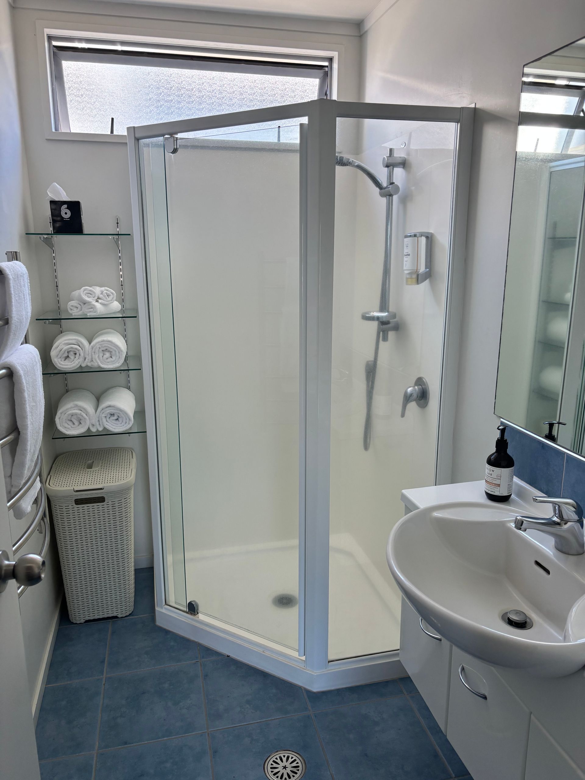 A bathroom with a corner shower stall, white walls, blue tiled floor, a vanity sink, and glass shelves holding rolled towels.