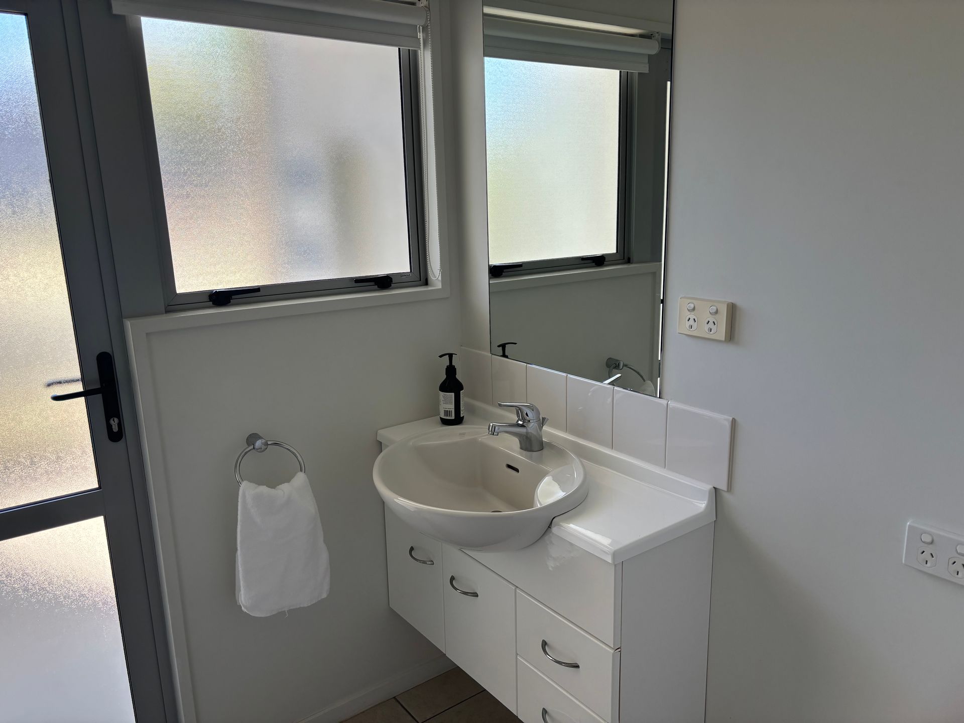 A white bathroom vanity with a basin sink, mirror, faucet, and a towel ring on a light wall next to a glass door.