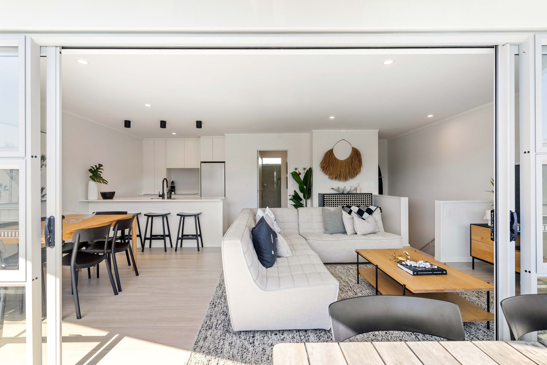 Bright living room with open sliding doors; white walls, sectional sofa, kitchen, dining area.