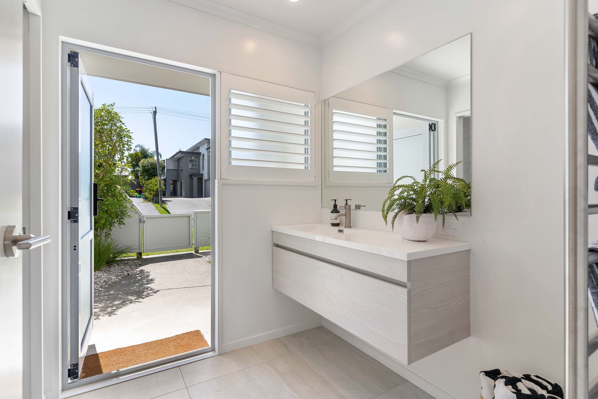 Bathroom with open door, floating vanity, large mirror, and shutters. Bright, white space with outdoor view.