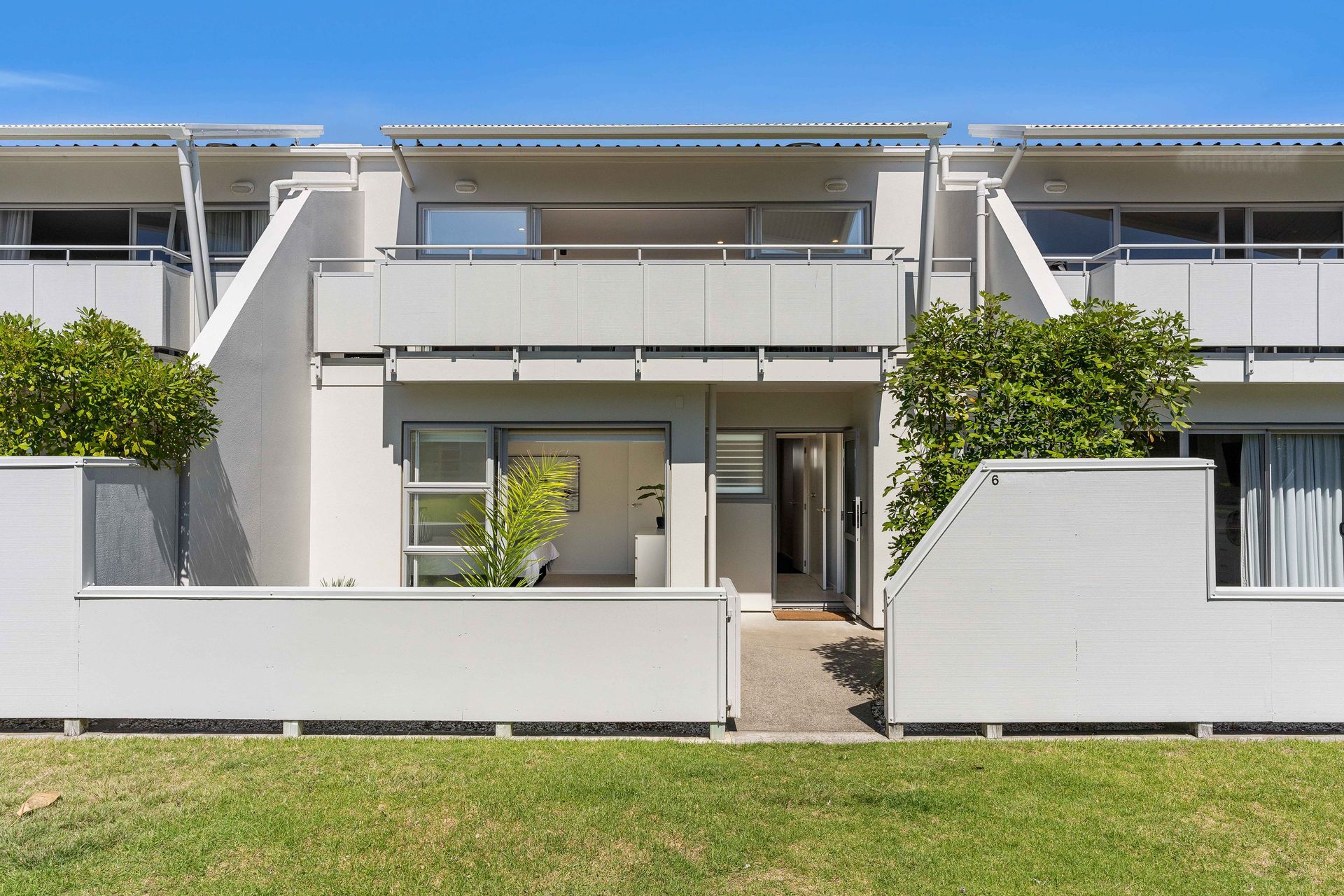White townhouses with balconies, trees, and a grassy lawn under a blue sky.