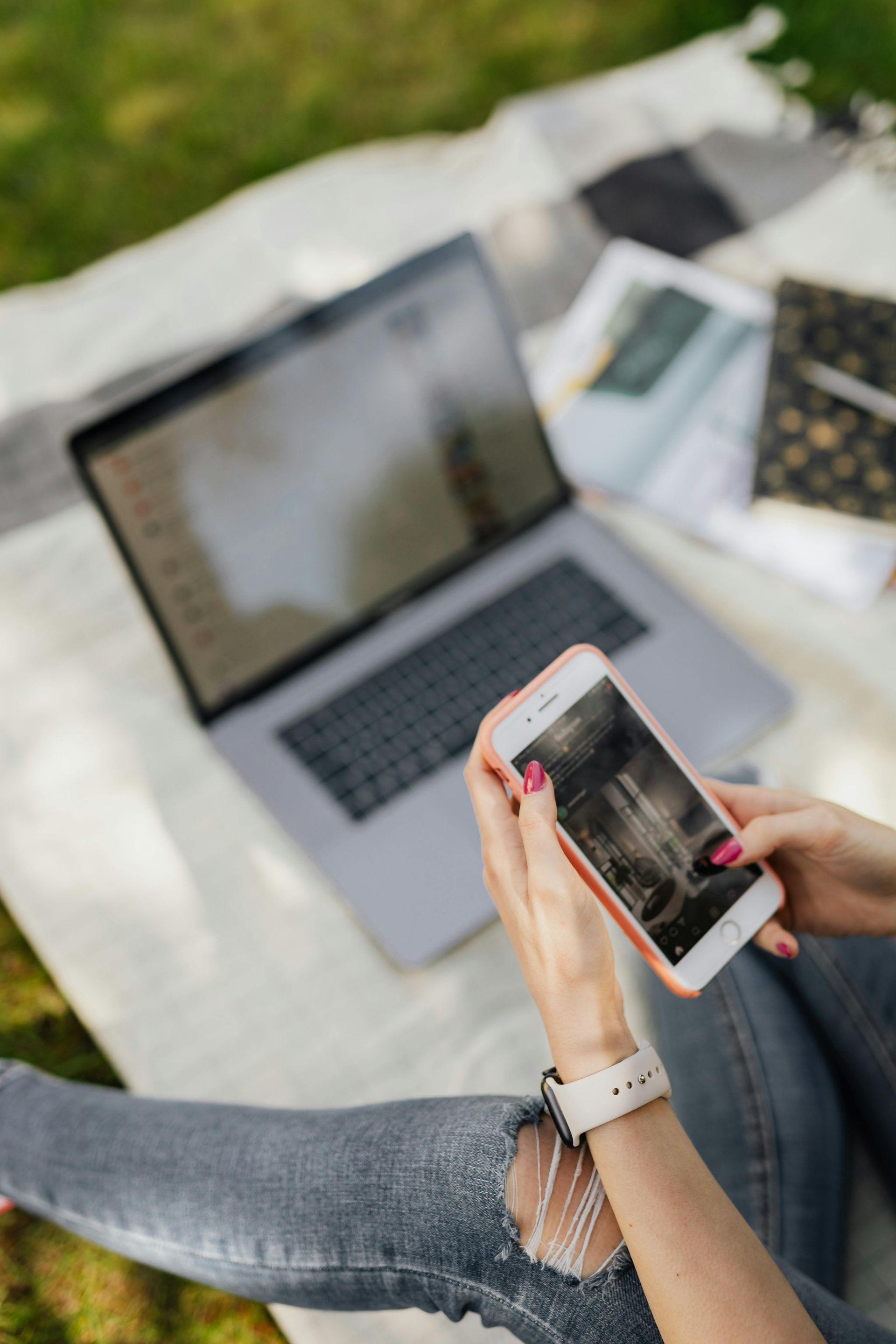A woman is sitting on a blanket using a laptop and a cell phone.