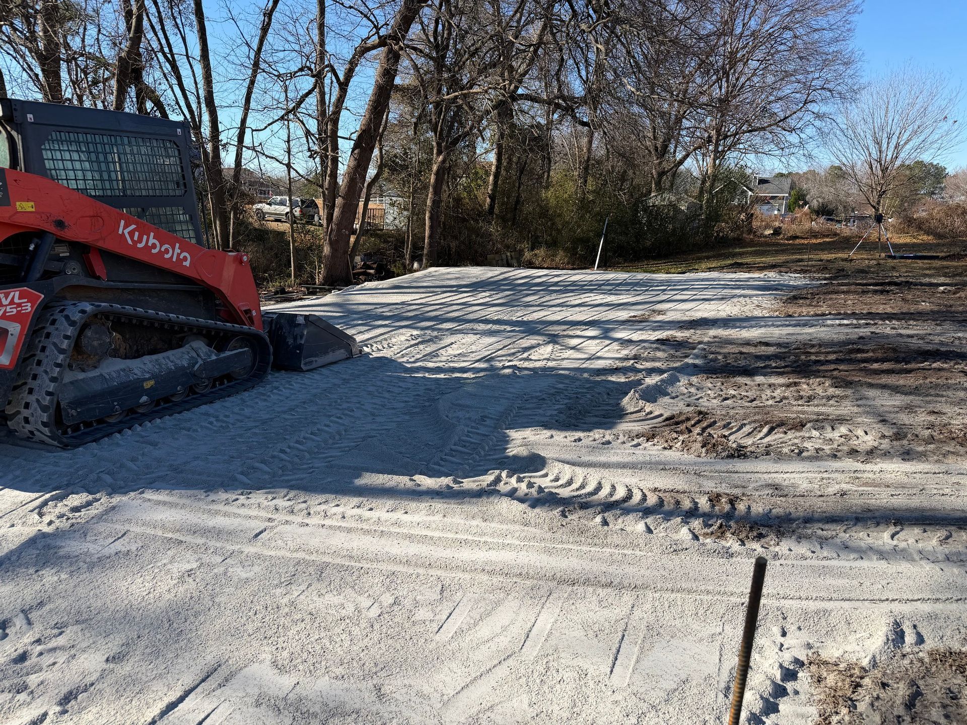 Red skid-steer clearing snow on a gravel driveway beside bare trees in winter.