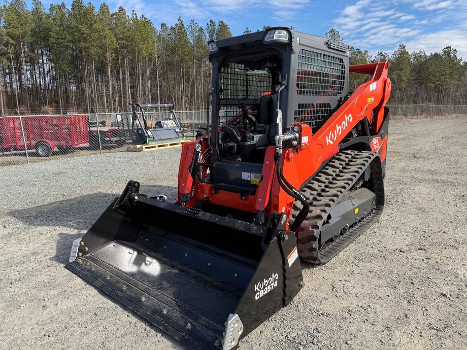 Red and black compact track loader with bucket on gravel lot, wooded background