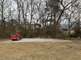 Small orange excavator on a gravel lot beside bare trees and brush.
