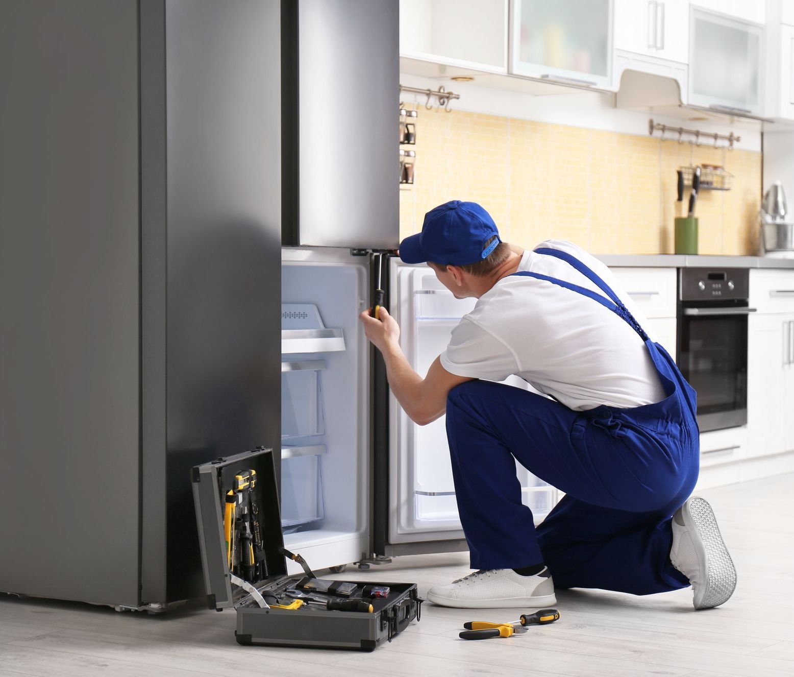 A male technician wearing a blue uniform and holding a screwdriver is repairing a refrigerator in a well-lit kitchen.