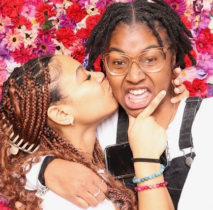 Woman kisses another woman’s cheek; they pose in front of a floral backdrop.
