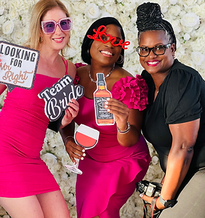 Three women in front of a flower wall holding photo props. Two wear pink dresses, smiling. One holds a camera.