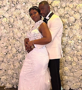 Newlyweds pose in front of a white floral wall. Bride in white gown, groom in tuxedo, embracing.