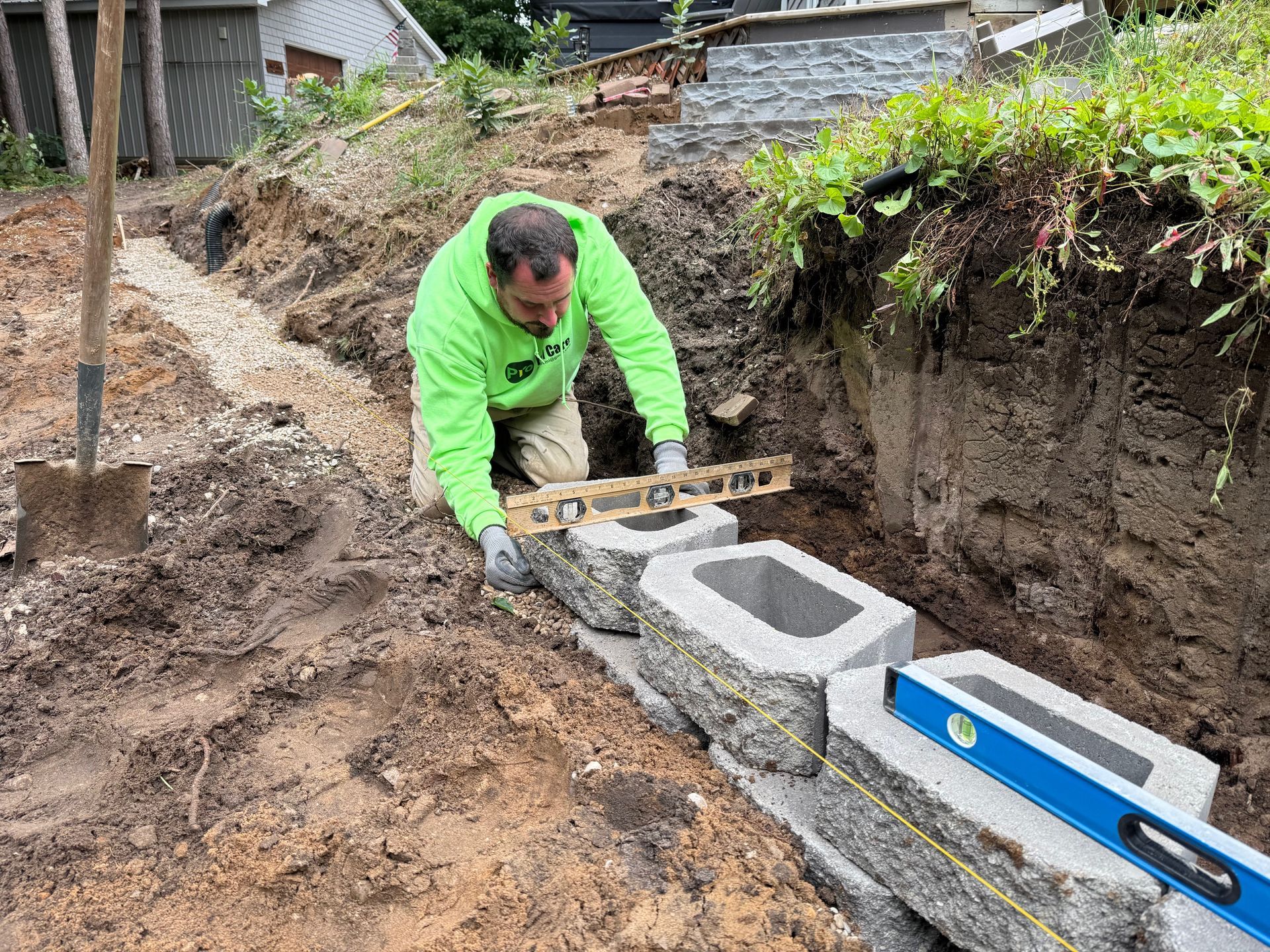 Man using a level to build a retaining wall with gray blocks in a dirt trench.