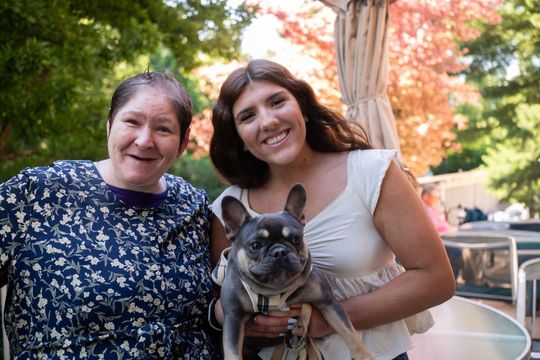 Two smiling women holding a French bulldog outdoors with autumn trees behind them