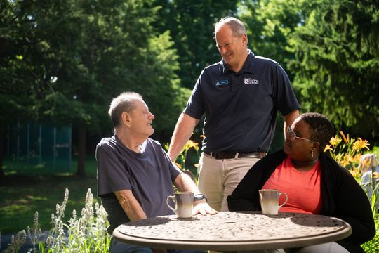 Three people chatting outdoors around a round table in a sunny garden