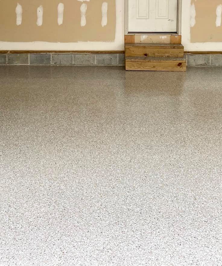 A garage floor with a smooth, light-colored, speckled epoxy finish next to a wall with drywall patches and wooden steps.
