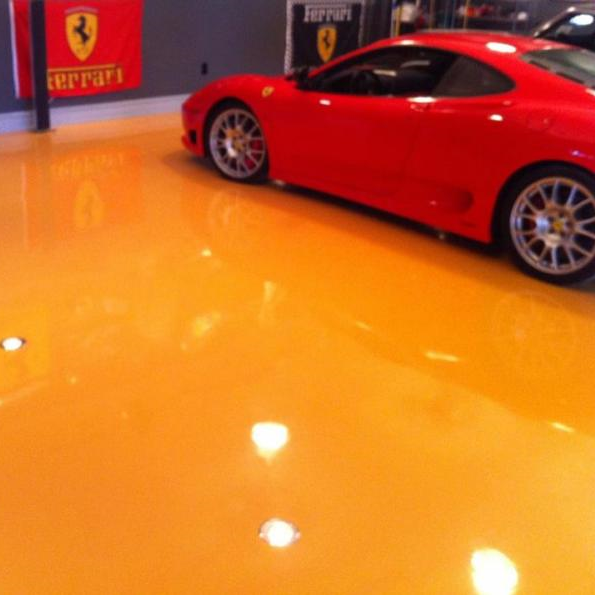 A bright red Ferrari sports car parked on a glossy, reflective orange epoxy garage floor with Ferrari banners on the wall.