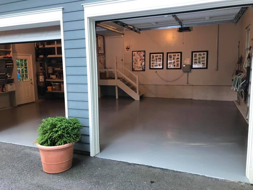 A two-car garage with gray epoxy floors, a white staircase, and three framed pictures hanging on the back wall.